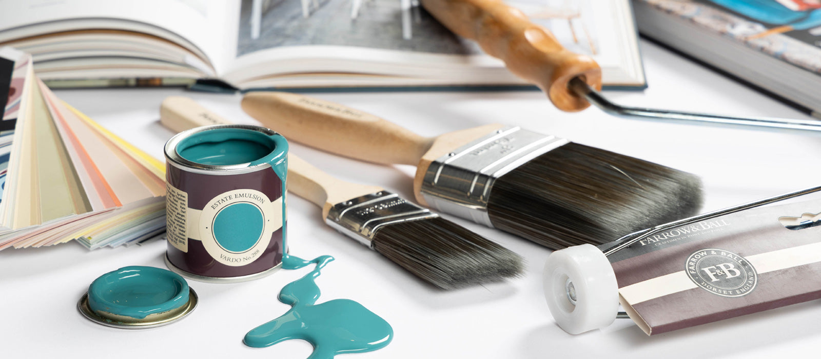 Übersichtlich aufgereiht von links ein Medium Colour Book, ein offener Sample Pot aus dem ein Farbbeispiel in Türkis gekippt wurde. Daneben liegen zwei Flachpinsel von Farrow and Ball und ein Steckbügel. Im Hintergrund erahnt liegen zwei Bücher