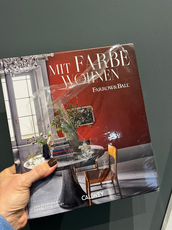 In der Hand gehalten, das Buch "Mit Farbe wohnen" im roten Cover auf dem ein runder Tisch mit einem Stuhl abgebildet ist.